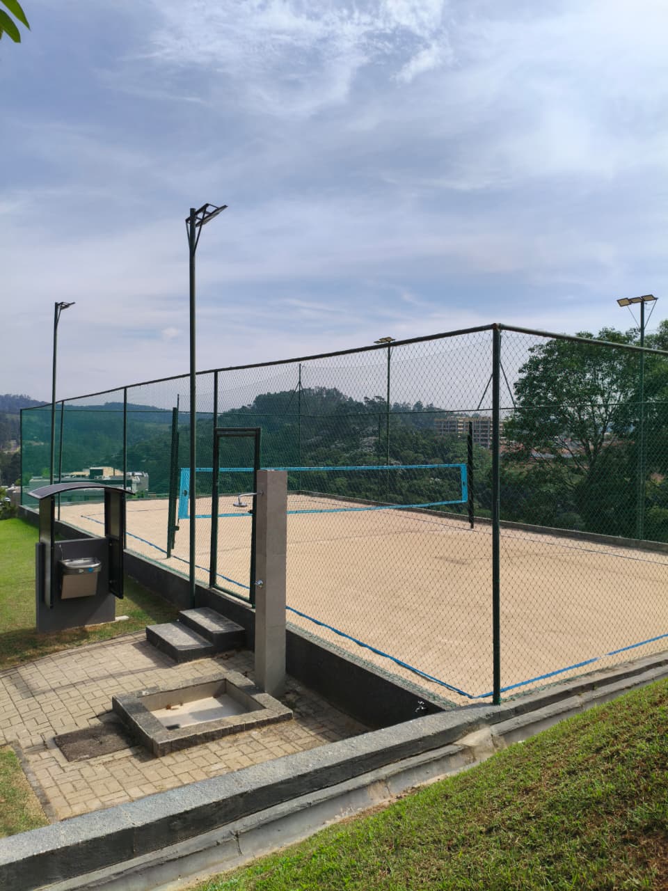 Lateral da quadra de beach tênis com bebedouro, placar e alambrado iluminado