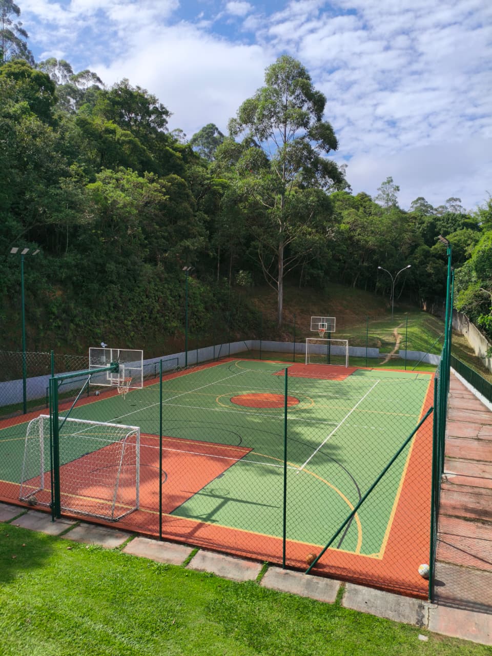 Quadra poliesportiva com piso verde e laranja, traves e cestas de basquete cercada pela mata nativa