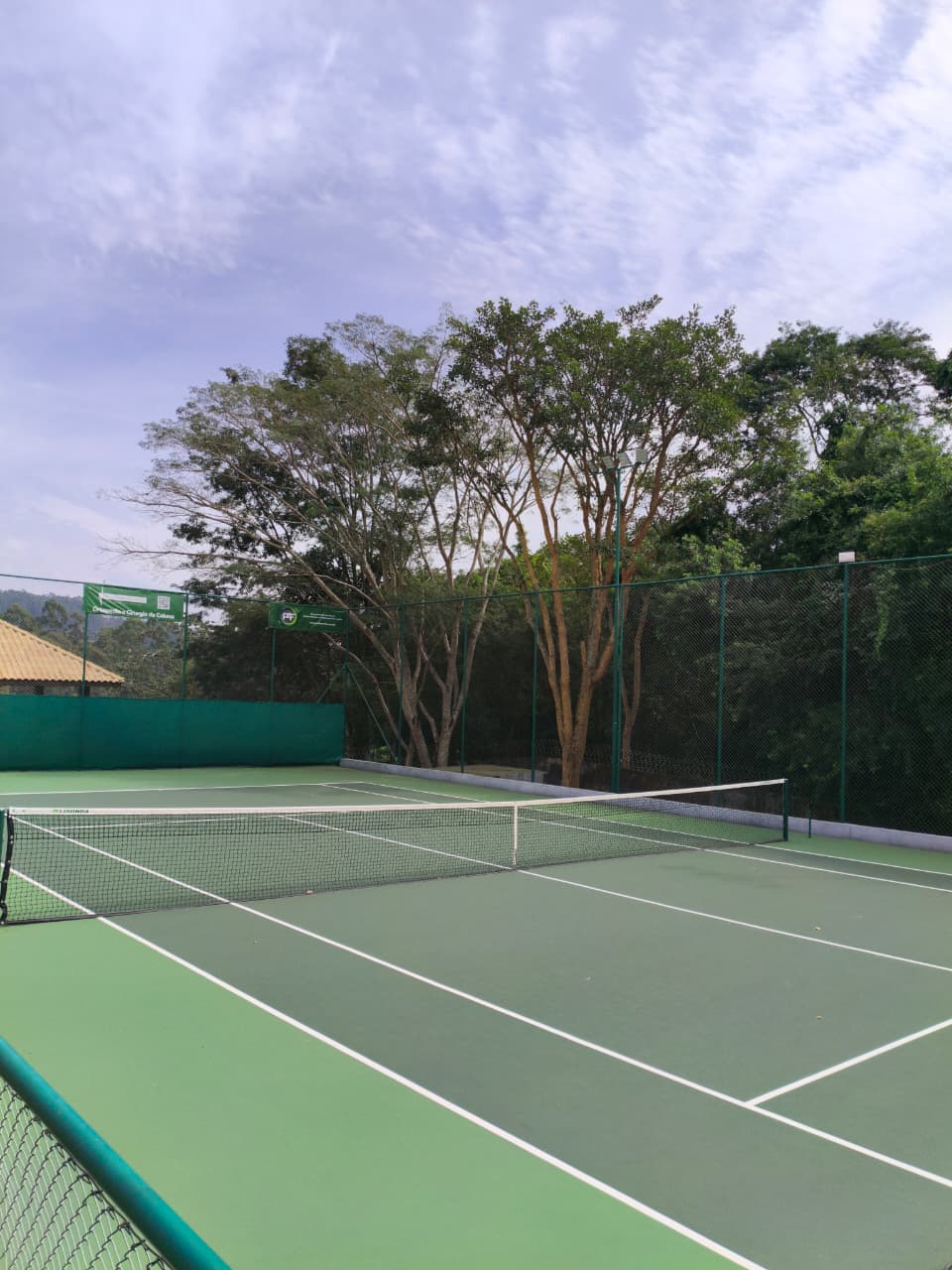 Vista em perspectiva da quadra de tênis com alambrado, refletores e árvores frondosas