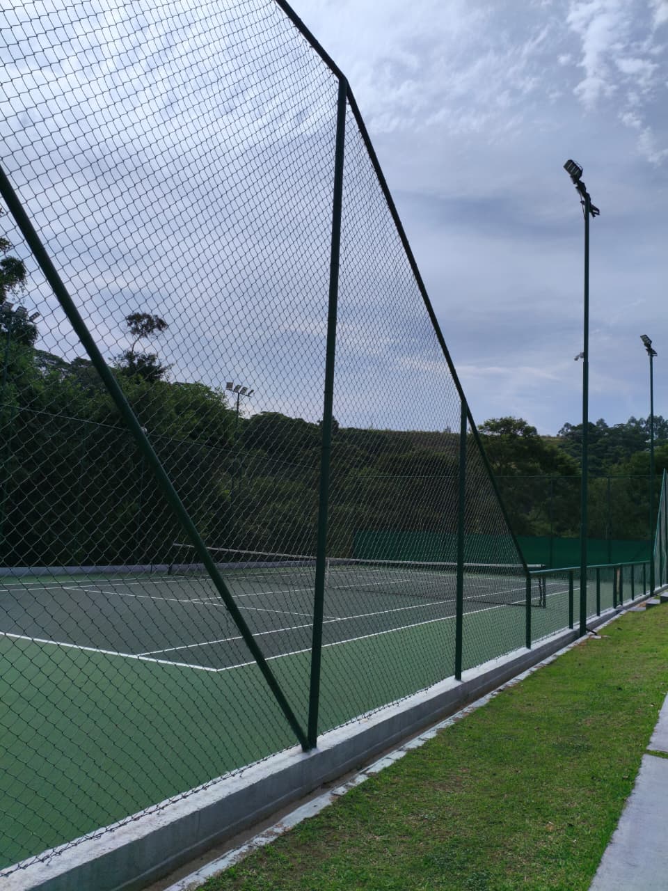 Lateral da quadra de tênis pelo alambrado, com encosta verde e refletores
